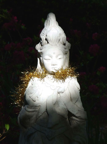 A serene stone statue draped with golden tinsel, illuminated by soft light, surrounded by vibrant flowers